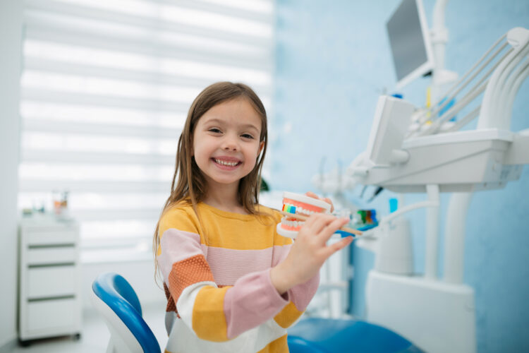 Portrait of a happy little girl sitting on a dentist chair and holding a model of human teeth and a tooth brush pretending to brush them, looking at camera